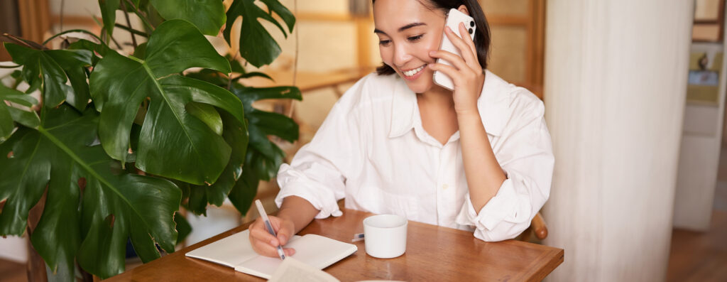 A woman sitting at a table, talking on a cell phone, while writing on paper in front of her.