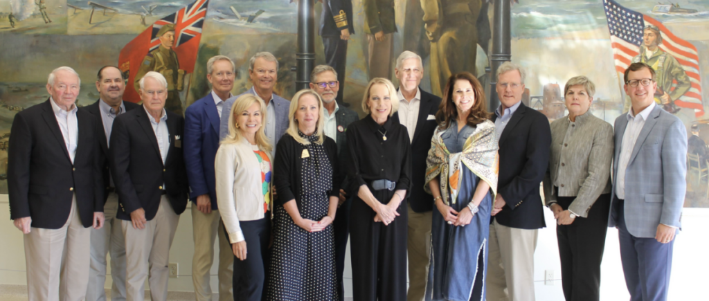 The image displays a group photo of nine people standing side by side in front of what appears to be an official backdrop featuring a portrait and a flag. The individuals are dressed in various styles, including jackets and dresses, suggesting an occasion or event of significance. They all seem to be posing for the photograph, with some looking directly at the camera while others are slightly turned or looking off to the side.