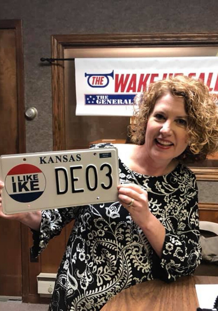 Woman holding license plate that reads ""KANSAS IKE DE03"".