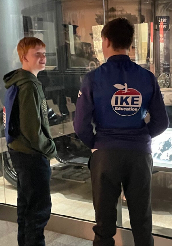 Two young individuals standing next to each other in front of a display case, both dressed casually; one wearing a blue jacket with 'IKE' printed on the back and the other in a green jacket with what appears to be a red and white logo.