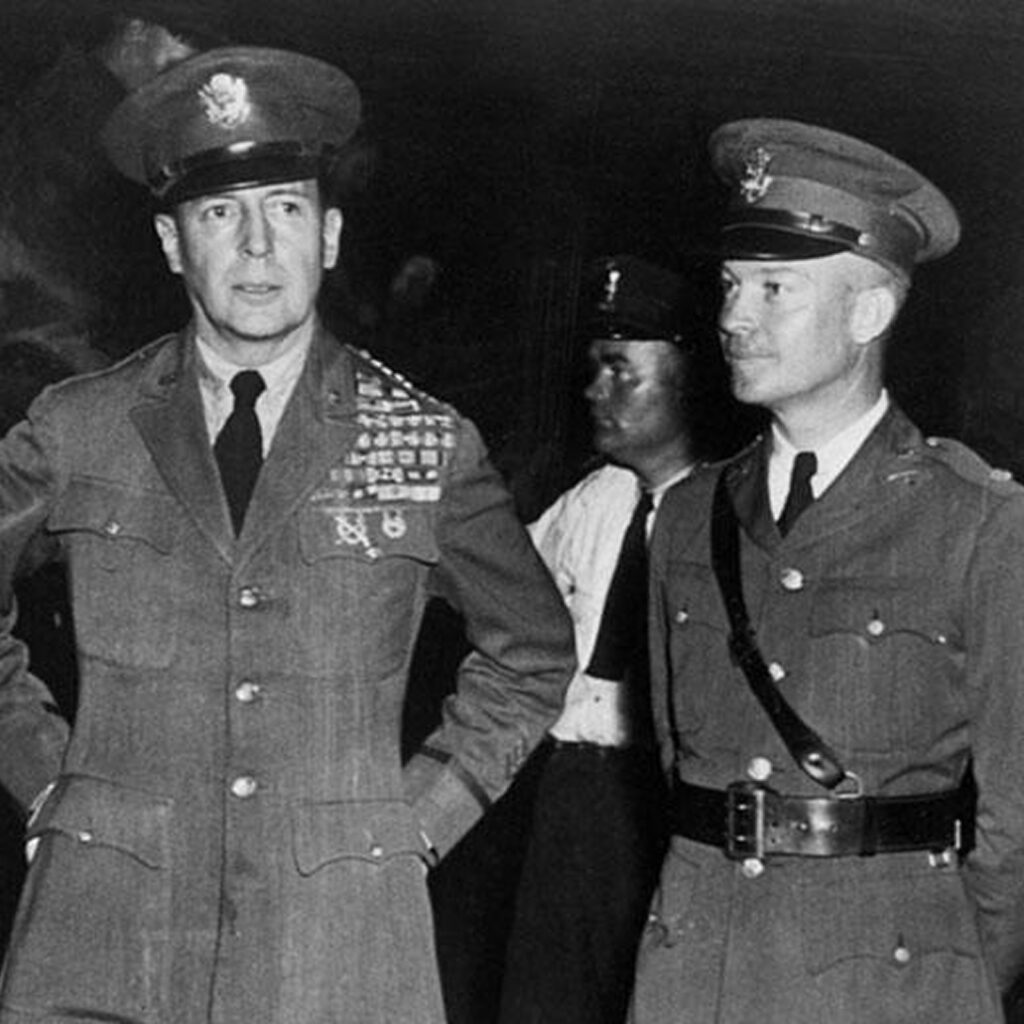 Three men in military uniforms, standing close together. All three wear medals on their chests. The man on the left has a tie and is looking forward. The other two men appear on the right; one has his hands behind his back, while the other seems to be pointing. They are standing against a dark background.