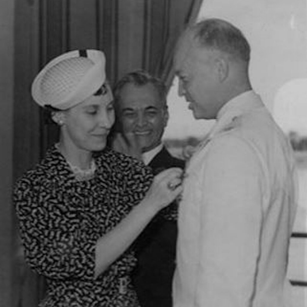 A woman in a vintage dress and hat is pinning a medal onto a man's jacket who is wearing a suit and tie. The man is looking down while smiling at the women, and a bald man stands slightly behind them, laughing.