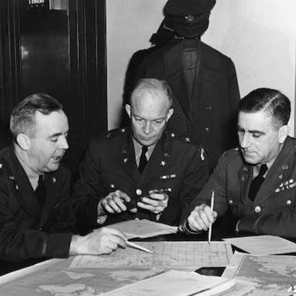 Three men in military attire are seated at a table, examining a map spread across its surface. The room appears to be an office setting with a formal ambiance. Two of the men are leaning over the document, holding a pen.
