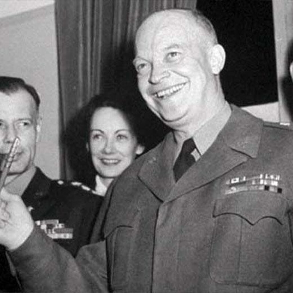 This is a black-and-white photograph featuring Dwight D. Eisenhower in military uniform. There are some other people standing around him. The setting appears to be indoors, possibly during a speech or award presentation.