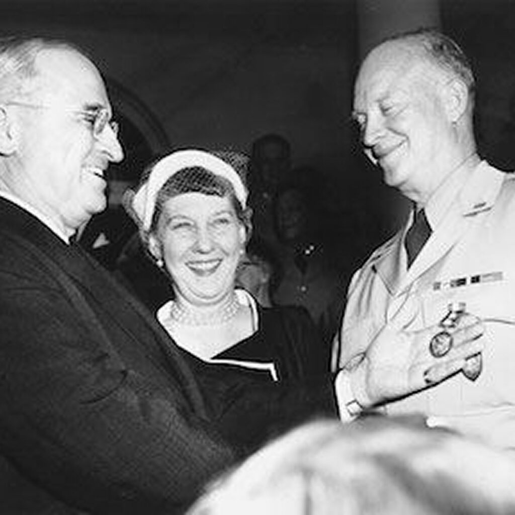 Two men and a woman smiling and standing close together in a social setting, smiling. The man on the right is wearing a tie and has a woman beside him, and the man on the left has a dark suit on; all three are dressed formally.