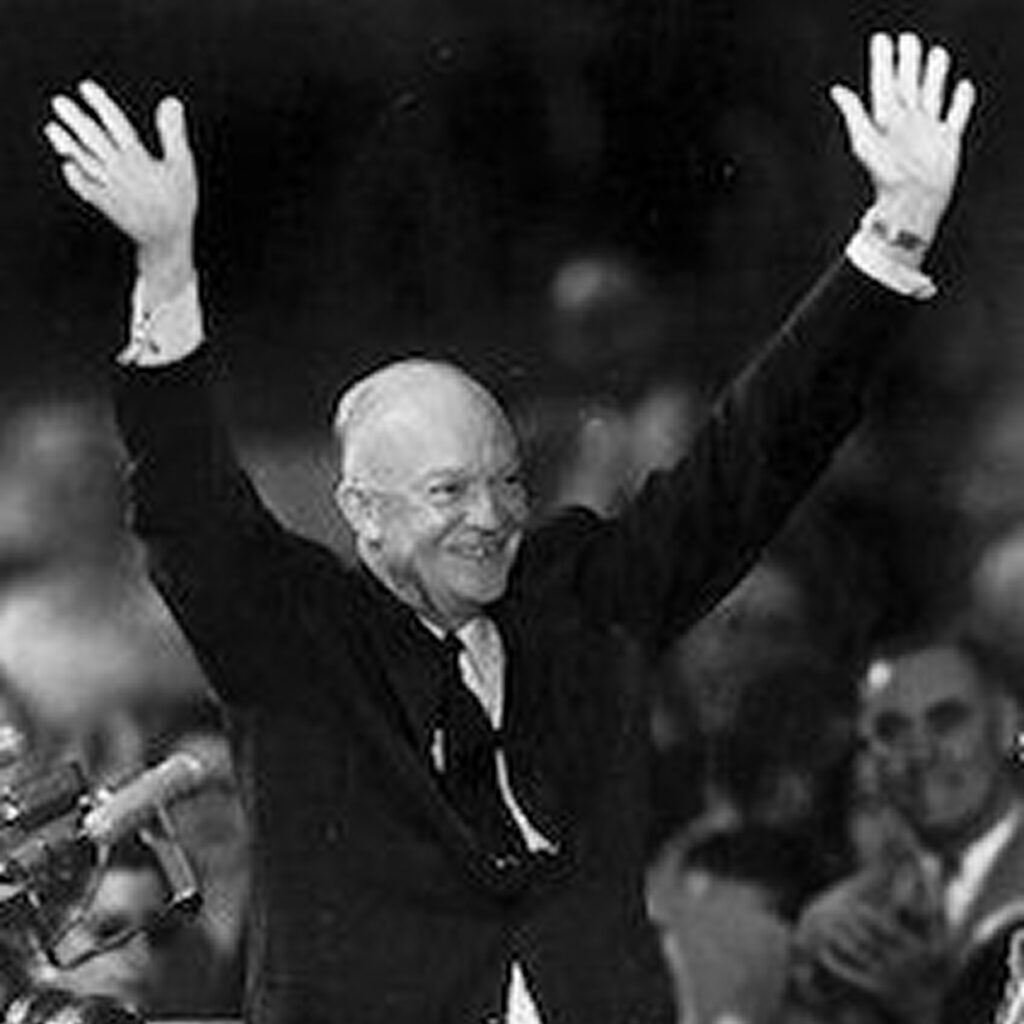 Dwight D. Eisenhower standing on stage at an event, raising his arms in the air and waving to the crowd.