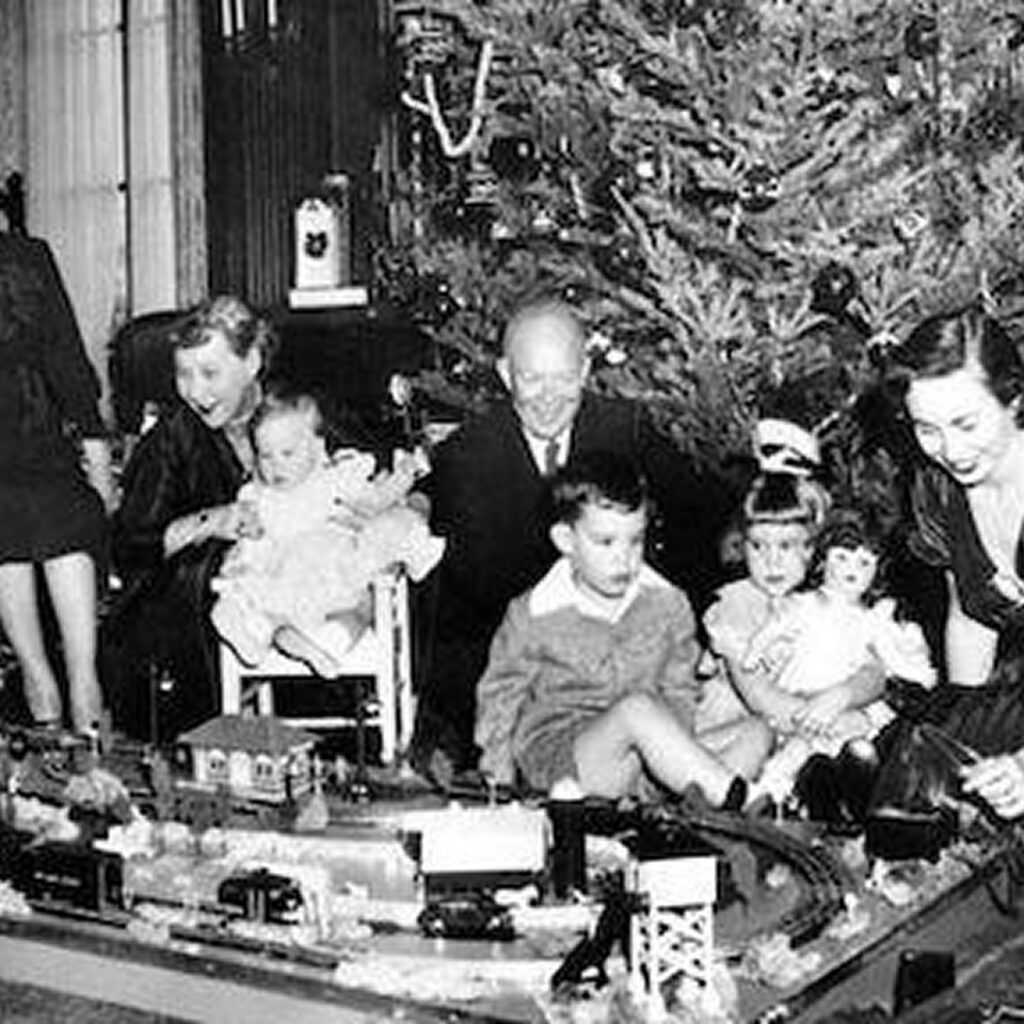 A group of people gathered around a toy train set. The room is decorated for Christmas with a tree in the background. The people are sitting and standing near the train display.
