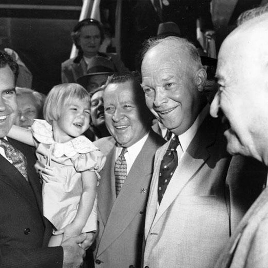 A black and white photograph capturing a moment with a group of people. Dwight D. Eisenhower is at the center, with several men and children around him. All are smiling.