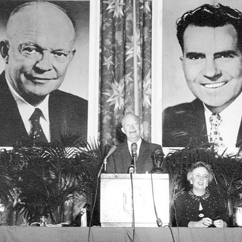 The image is of an indoor event, likely a political speech or panel discussion, where two individuals are seated at a podium in front of banners featuring portraits of men.