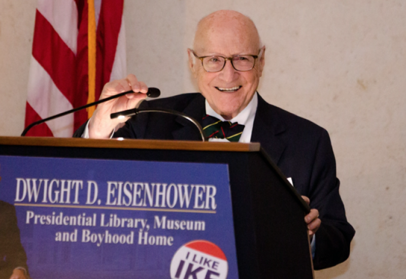 Elderly man speaking at a podium beside American flags.
