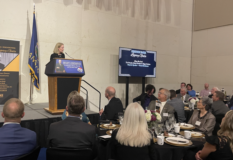 A woman speaking at an event in front of a podium and attendees seated around tables with place settings, wine glasses, and presentation screens displaying images and text.