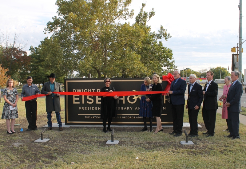 A group of people gathered at an outdoor event, cutting a ribbon in front of a sign saying "Dwight D. Eisenhower Presidential Library & Museum."