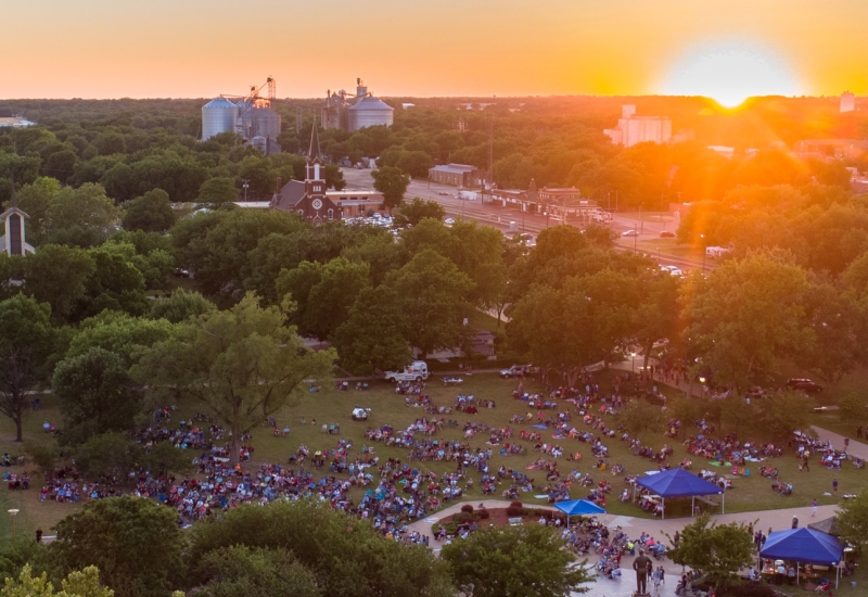 The image captures a vibrant scene at sunset. A crowd of people is gathered in a park, enjoying the spectacle under a clear sky with hues of orange and blue blending into the horizon. The silhouette of trees forms an attractive backdrop to the gathering. In the background, there's a building visible and on the left side, a car can be seen parked. On the right side, there's a water tower standing tall. The overall atmosphere is one of leisure and communal enjoyment.