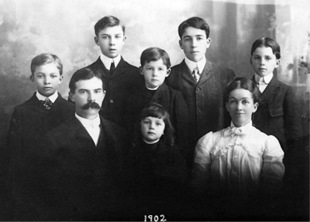 Family portrait from the early 1900s with 8 individuals in formal attire. The group includes adults and children, some of whom are seated. The style of clothing indicates an older time period.