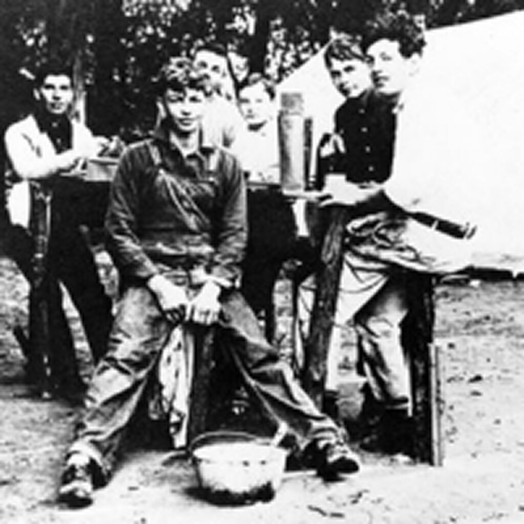 A black and white vintage photograph captures a group of young men posing outdoors at a picnic table. They all wear casual attire appropriate for outdoor activities from that era.