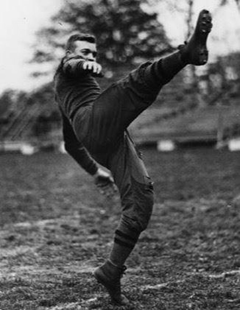 A man in old football uniform kicking ball on field.