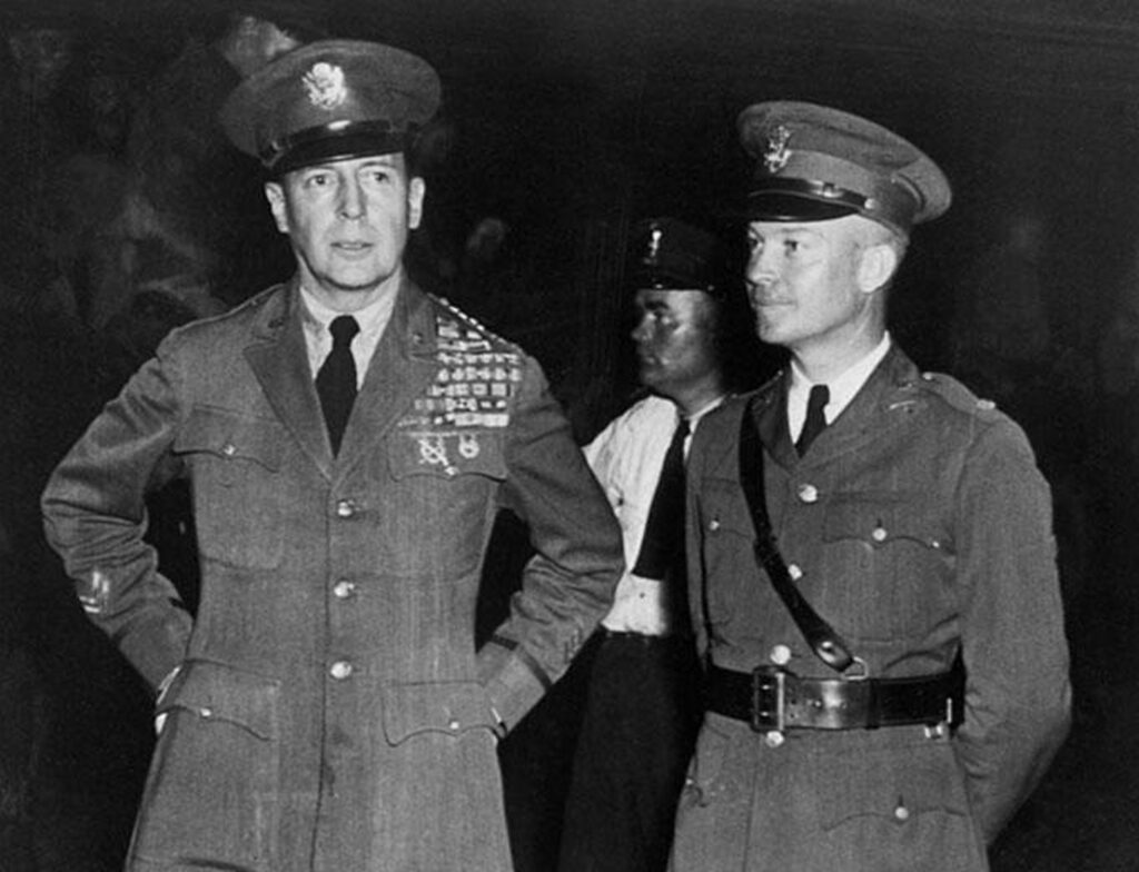 Two men in military uniforms standing next to each other. The man on the left has decorations pinned to his shoulder and is looking towards his right with his hands behind him. The man on the right is wearing a similar uniform, also decorated with medals, and is also looking off to the side.
