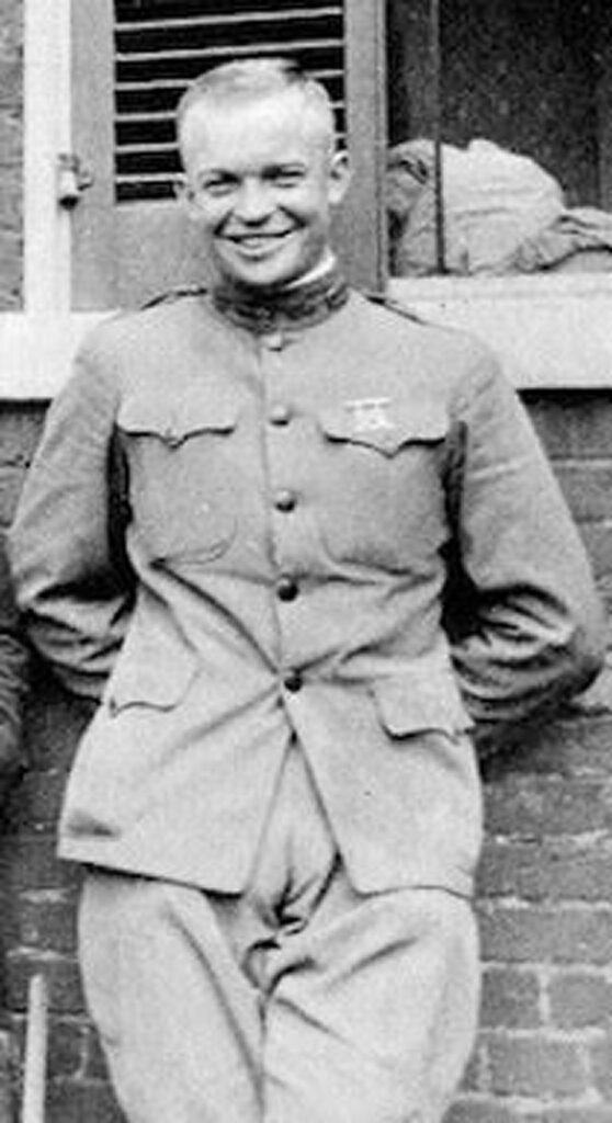 A young Dwight D. Eisenhower in military uniform leaning against brick wall. He is smiling at the camera.