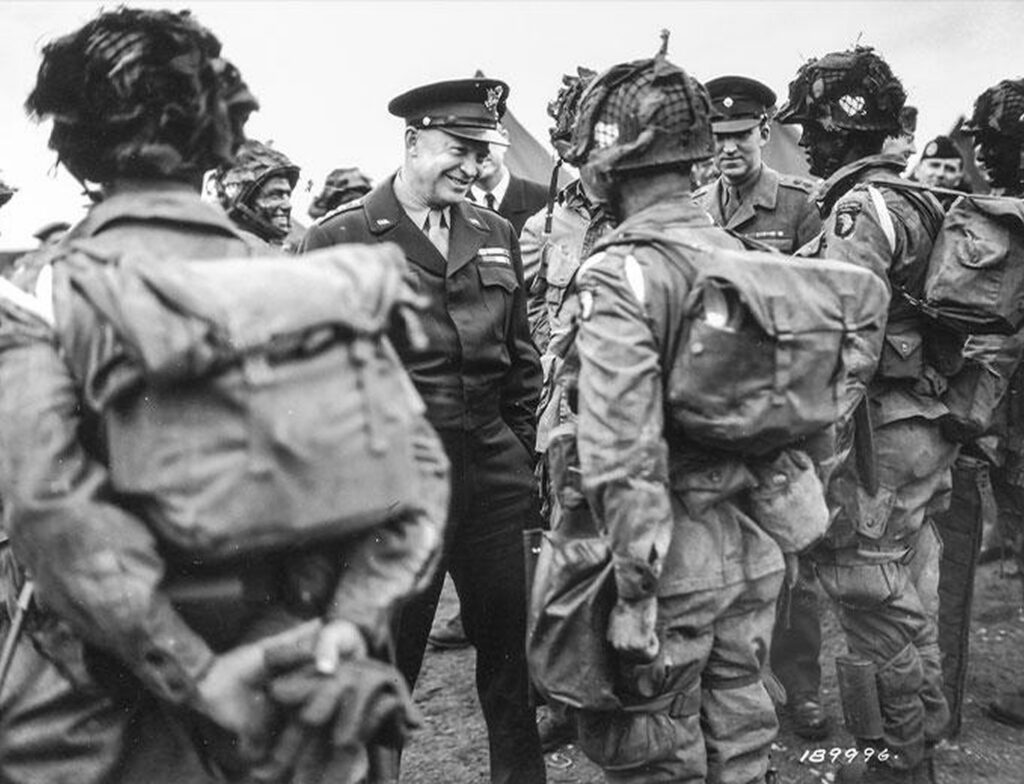 A group of military men in uniform standing near each other, with Dwight D. Eisenhower seeming to be smiling at them and conversing with them at the center.