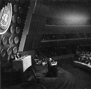 The photo appears to be from a political event inside a large stadium with a crowd seated in rows. There's a central stage where the main speaker is standing, addressing an audience. Above the stage, a large circular emblem with the letters "UN" prominently displayed on it.