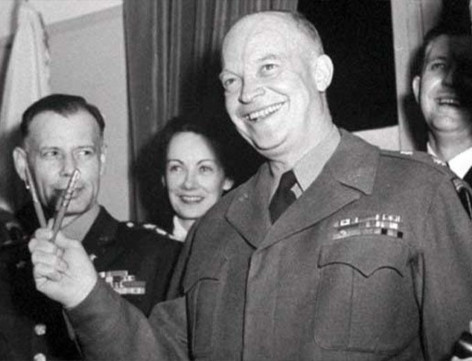 General Eisenhower holds two pens in a "V", smiling with men and a woman on Victory in Europe Day.