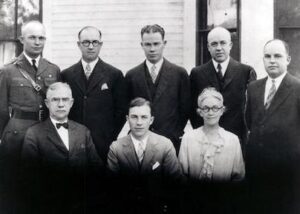 A family portrait of a group of men and one woman posing for a photograph; they are dressed in suits with visible ties. The setting appears to be a formal occasion.