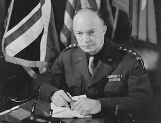 The image shows a black and white photograph of Dwight D. Eisenhower at a desk with flags in the background. He is dressed in formal attire and is writing on paper with a pen.