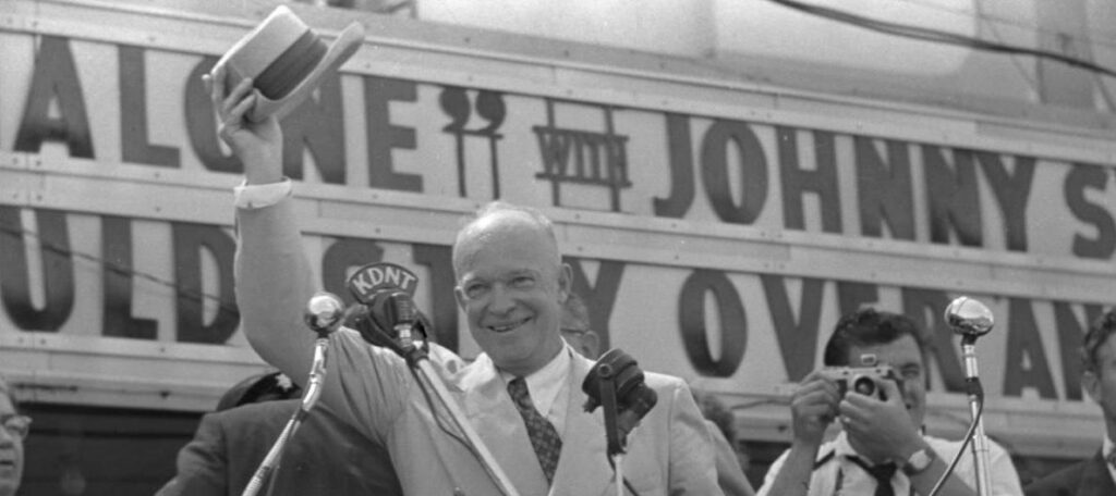 Black-and-white image of two people beside a campaign sign.
