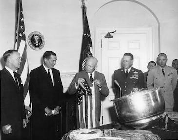 A black and white photograph showing a group of men in suits standing near the Oval Office door, with an American flag hanging behind them. The scene suggests it might be a meeting or event taking place within the White House.