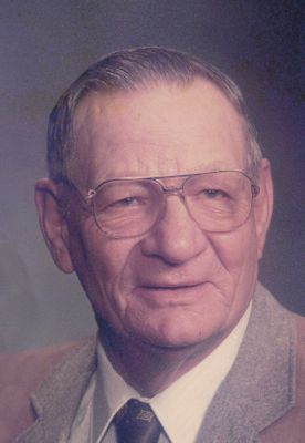 The image shows an older man with gray hair, wearing glasses and a dark tie, posing for a photo in front of a beige wall. He is smiling and looking directly at the camera.