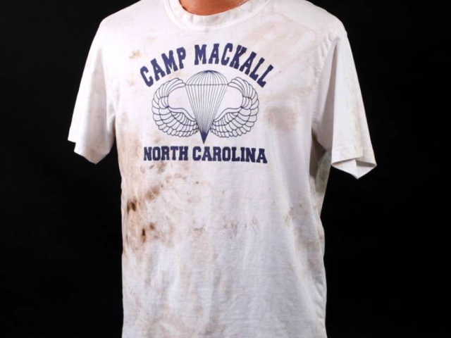 White souvenir shirt printed with “Camp Mackall, North Carolina” and an airborne insignia.