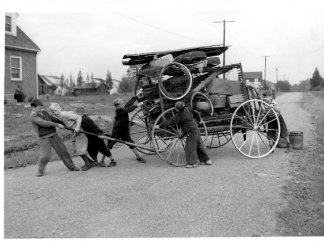 Children gathering scrap metal beside a wheeled cart during a wartime collection drive.