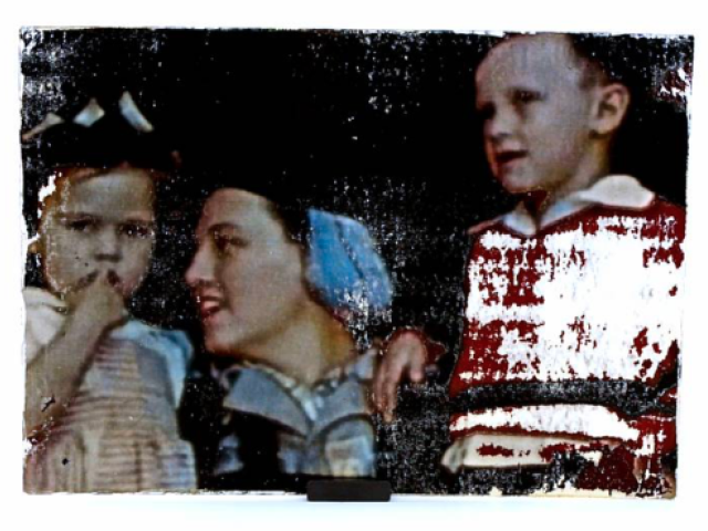Woman wearing a blue headscarf stands with two children beside a red-and-white bag.
