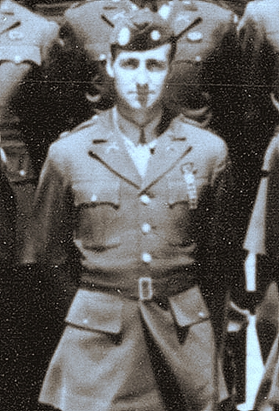 A group of military personnel in uniform, standing together. There is one man most visible in the center, wearing a hat and uniform with various emblems on it.