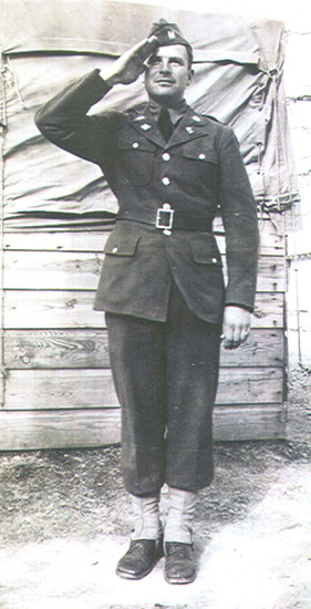 Man in military uniform saluting camera; he wears a hat and stands before a wooden structure with tarp covering it.