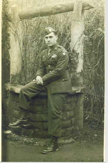 An old, sepia-toned photograph of a soldier sitting on a stone well for a picture.