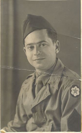 An aged photograph captures a man in military uniform. He is wearing a cap, and had an emblem patch on his shoudler. The man has dark hair and is smiling at the camera.