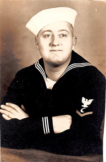 A sailor in military uniform with crossed arms, standing against a beige background.