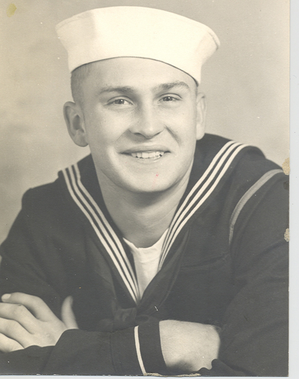 A black-and-white photo of a person dressed in a sailor suit, posing for a portrait. The individual is wearing a cap, and he is smiling at the camera.