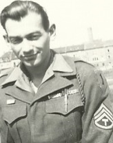 A man in military uniform smiling at the camera, with medals on chest and an insignia on his shoulder, standing outside.