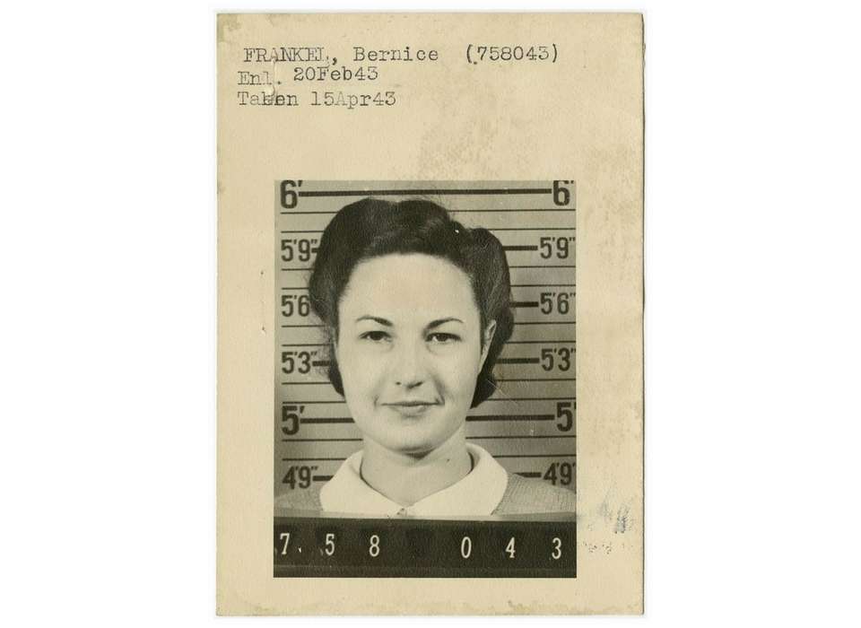 This is an old mugshot featuring a woman with short hair, wearing a collared shirt. She is looking directly at the camera. To the right of her headshot is a set of measurements with numbers next to each category.