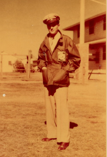 Old sepia-tone photograph of a male in military uniform standing outdoors, posing with hands in his pockets while standing outside.