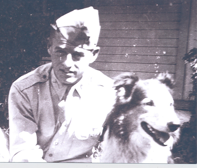 Sailor sitting outdoors in a white naval uniform and cap next to a dog.