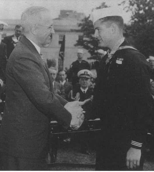 The image is a black-and-white photograph featuring two men shaking hands. The man on the right is wearing an military uniform with badges, while the man on the left is dressed in formal civilian clothing.