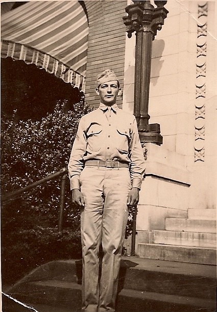 A sepia-toned photograph of a man in military uniform standing outdoors in front of a building. He has his hands at his sides.