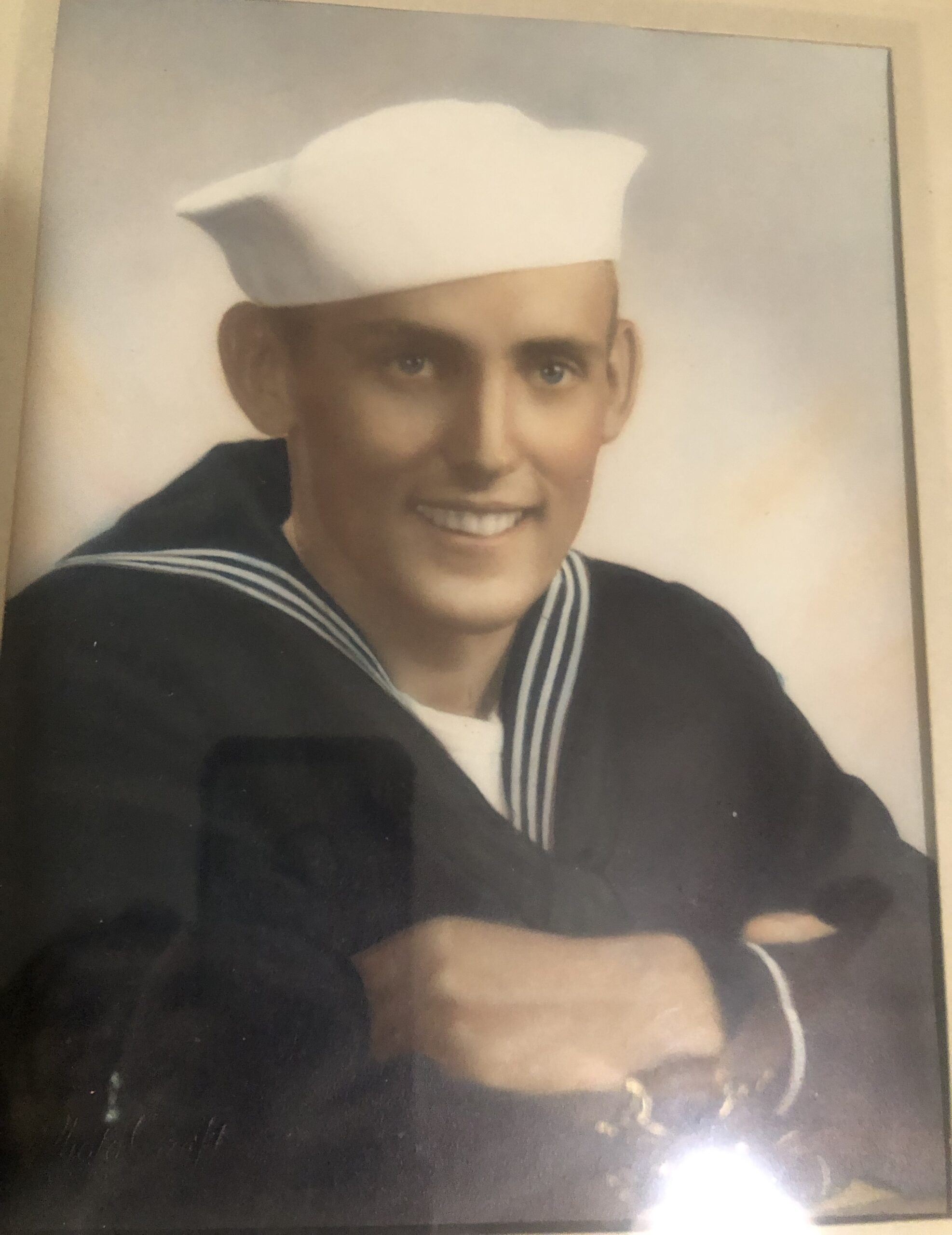 A sepia photograph featuring an older male in a U.S. Navy uniform. He is wearing a sailor's hat, and his uniform is complete with a peacoat and a tie. The man appears to be smiling slightly at the camera.