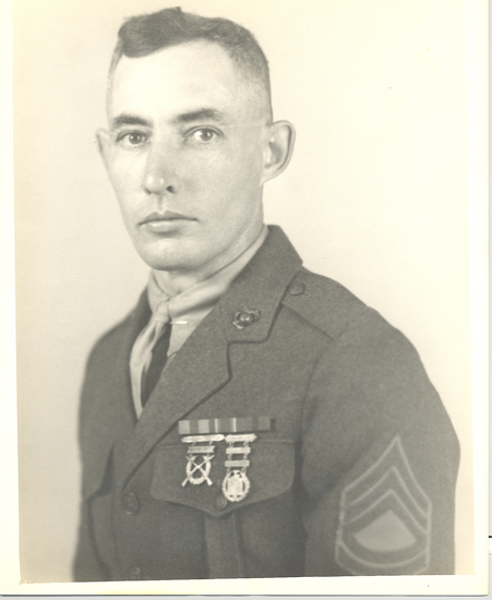 Old black-and-white photo of a soldier wearing a military uniform with ribbons and medals, posing for a portrait. He is standing upright, looking slightly to his right. The man has short hair and appears stern or serious in expression.