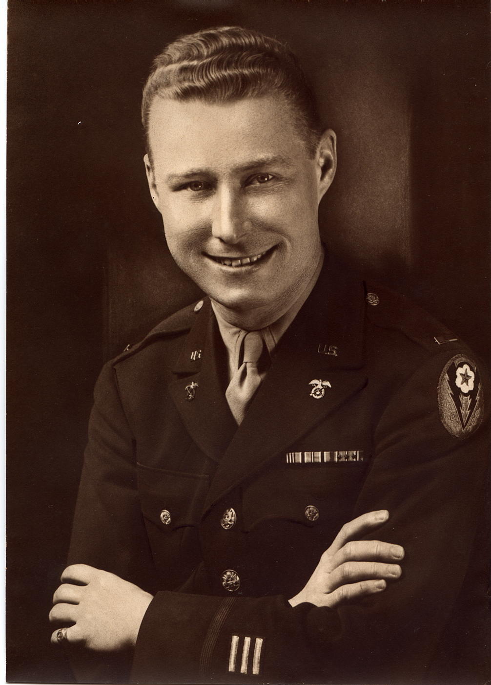 A sepia-toned photograph of a man in military uniform. He is smiling with crossed arms.