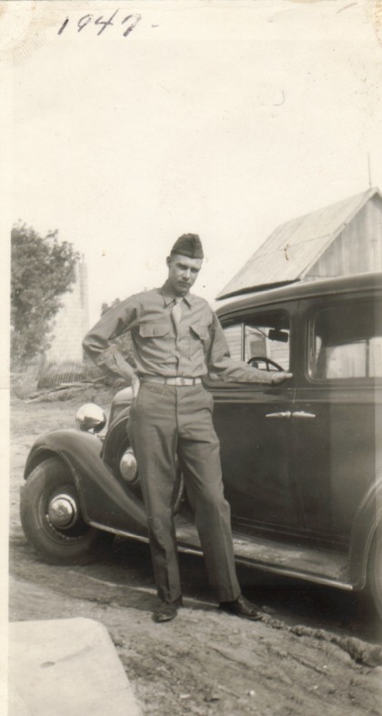 Black and white photo of an American soldier standing beside his vintage car, with a military uniform and cap.
