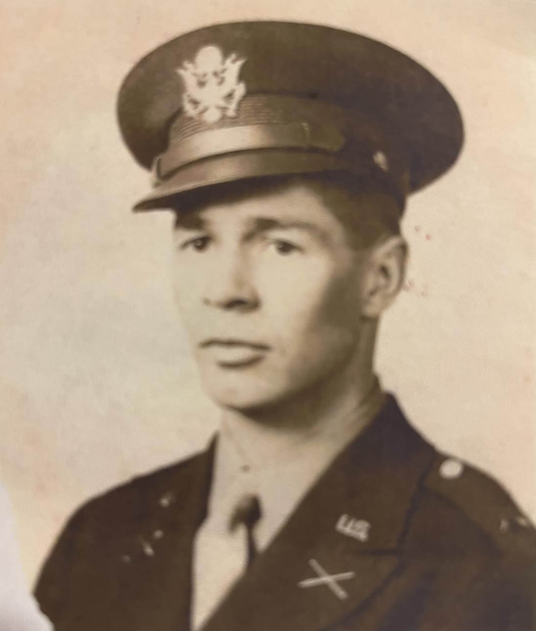 A man in military uniform, featuring a tie and badges, wearing an army cap with insignia, posing for a portrait.